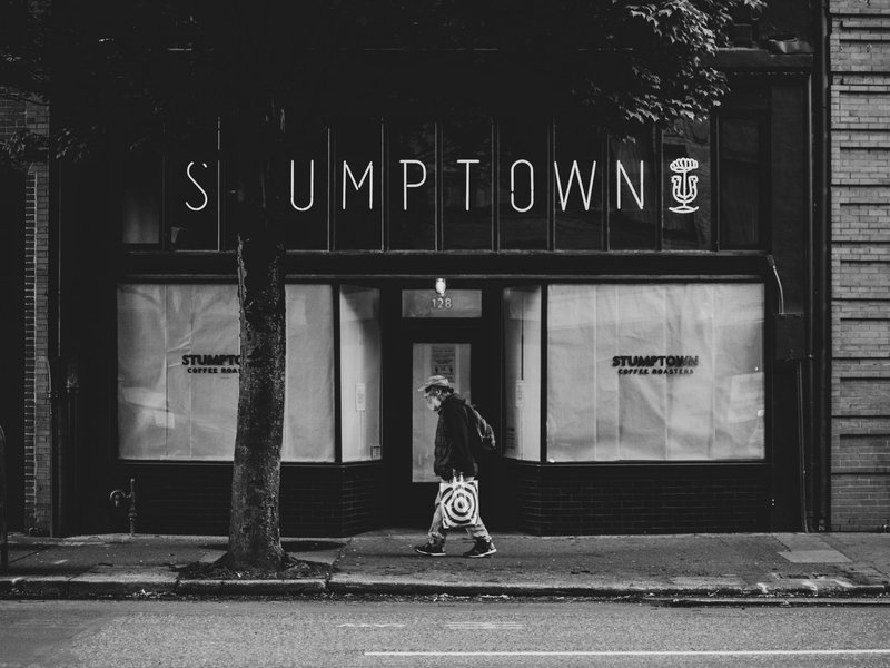 Black and white photo of a man with a shopping bag walking past a closed “STUMPTOWN” coffee shop
