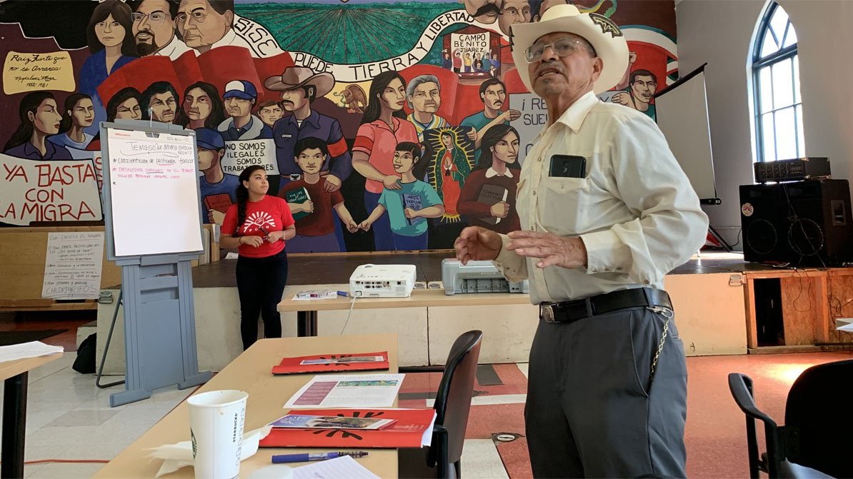 A Latino man in a cowboy hat speaking in front of a mural