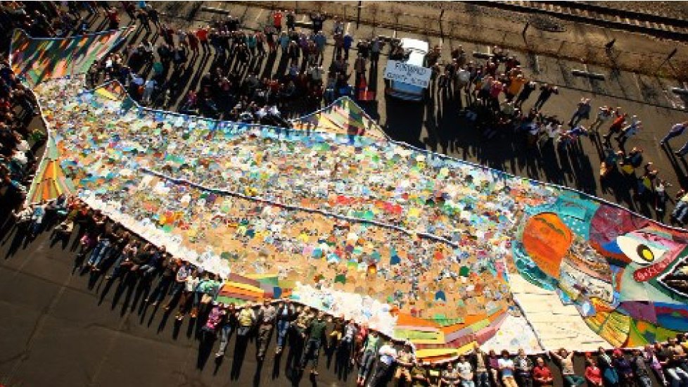 Overhead view of a sit in or other collective action, with ~100 people surrounding a large salmon artwork