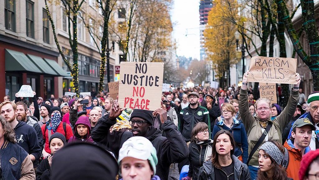 Group at a rally/protest. One person prominently holds a sign reading “NO JUSTICE NO PEACE”